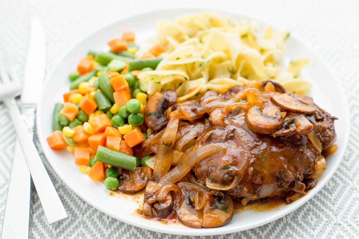 Salisbury Steak with Mushroom Gravy served with noodles and veggies on a round plate with veggies and butter noodles