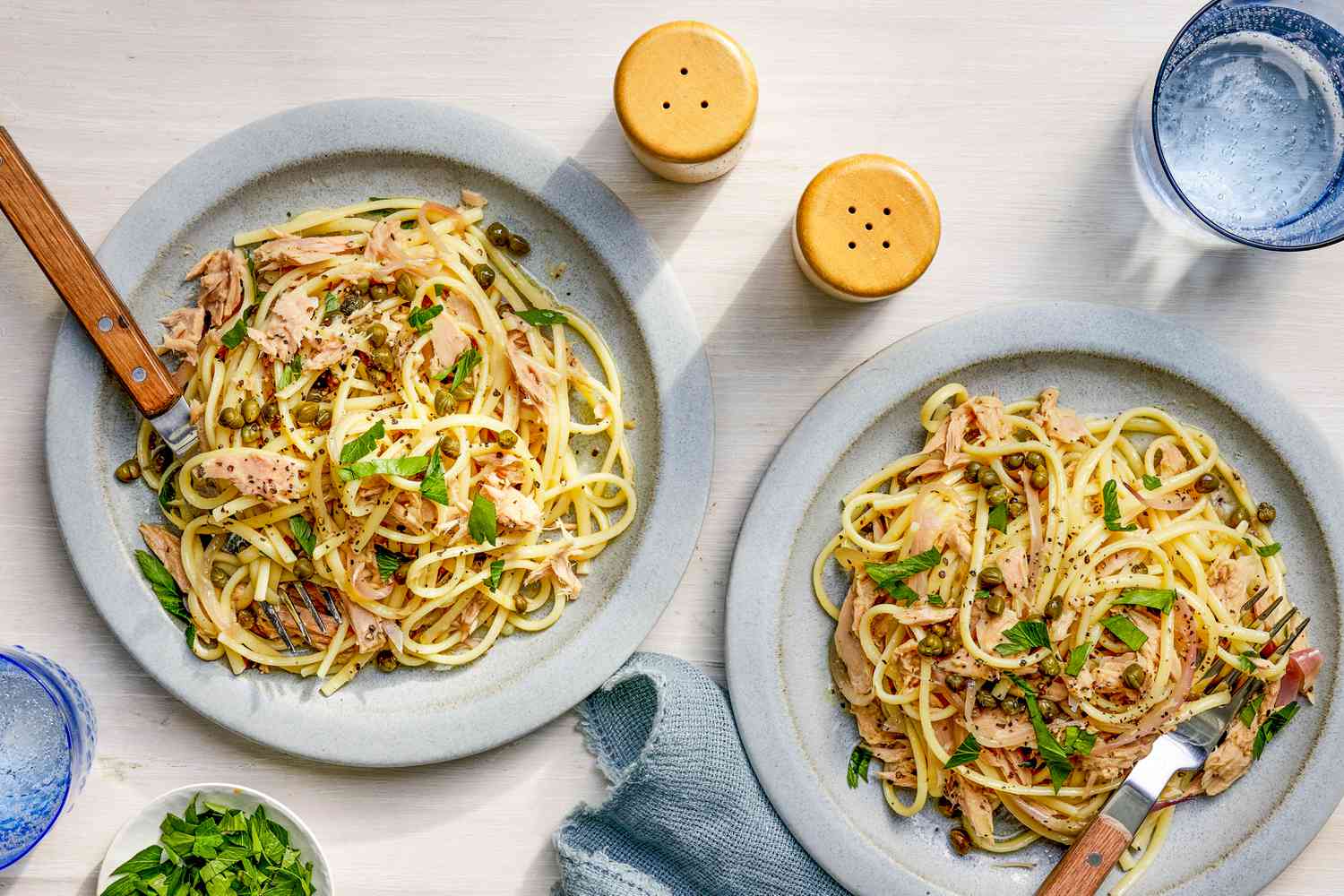 Linguine with tuna and capers on two plates at a table setting with a bowl of parsley, a denim table napkin, salt and pepper shakers, and a glass of water