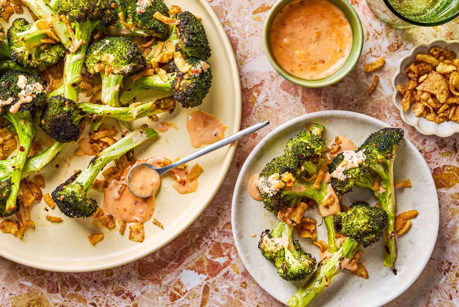 large plate of Bang Bang Broccoli smothered with Bang Bang sauce, a cup of the sauce on the side and spoon of sauce on the plate, small portion of the dish on a small plate, all garnished with fried onions, small dish of fried onions on the side
