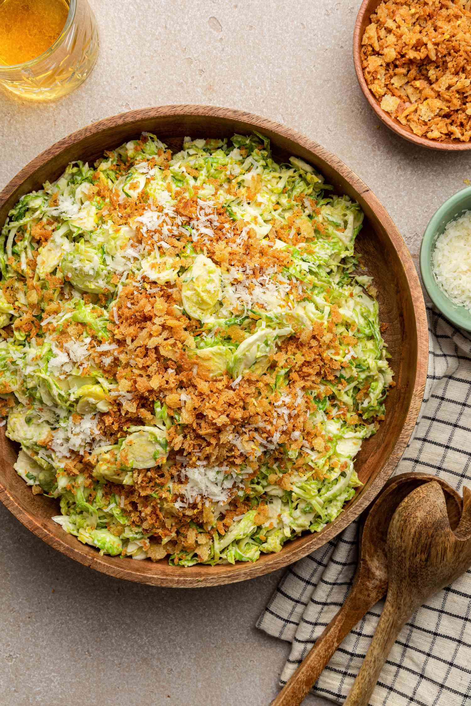 Bowl of Brussels Sprouts Caesar Salad with Lemony Breadcrumbs Next to Serving Utensils on a Table Napkin, a Bowl with More Toppings, a Bowl with Shredded Parmesan, and a Glass
