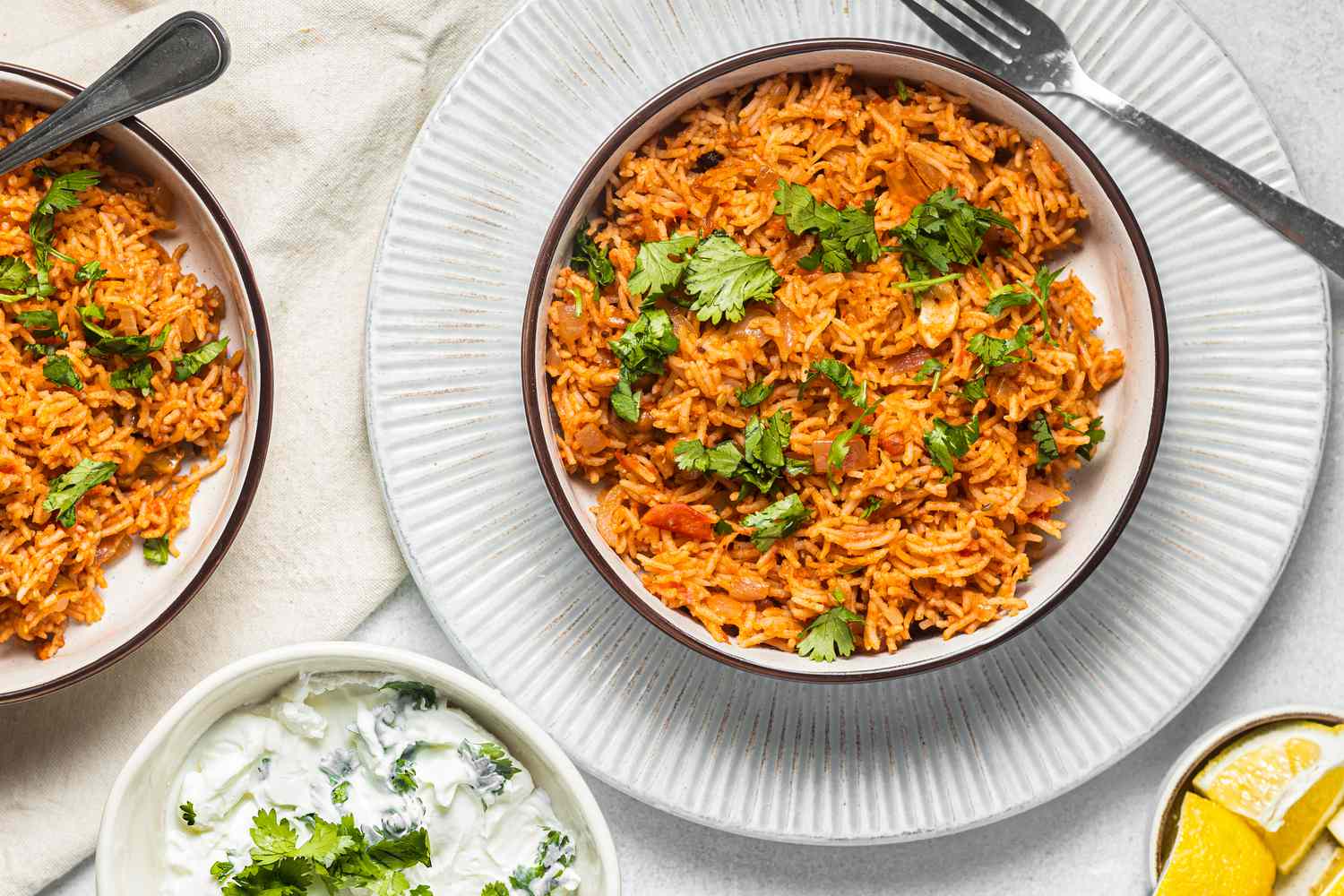 Tomato Rice with Coriander Leaves Next to a Bowl of Raita, a Bowl of Lemons, and Another Bowl of Tomato Biryani