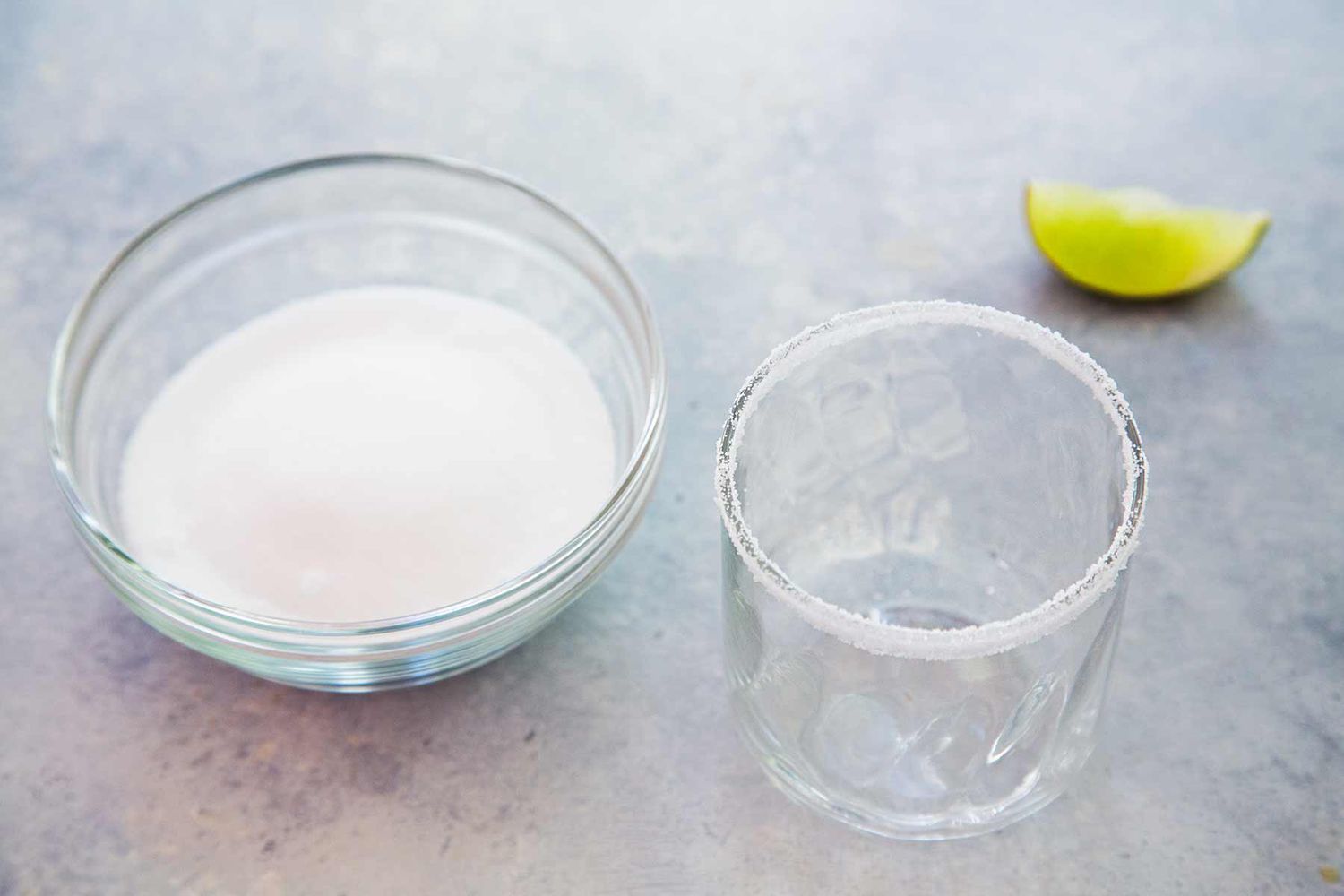 A bowl of salt, an empty glass with salt on the rim, and a lime wedge in the background 