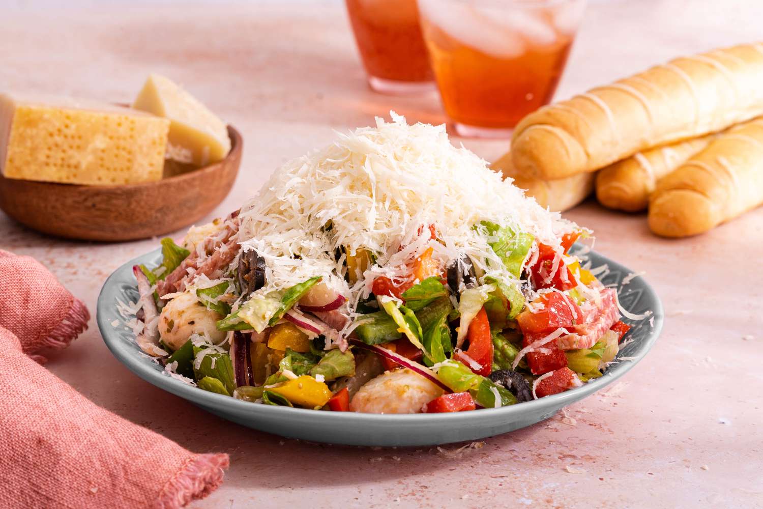 Plate of Pizza Salad Topped With Grated Parmesan, and in the Background, a Pink Kitchen Towel, a Bowl With Small Parmesan Blocks, Breadsticks, and Two Iced Drinks