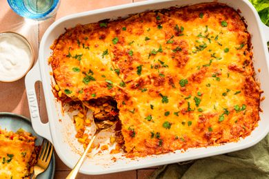 Chicken taco casserole (with a serving cut out) at a table setting with glasses, a serving on a plate, a side salad, a bowl of sour cream, and a kitchen towel