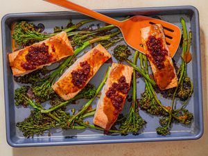 Overhead view of a baking sheet of four cooked salmon filets topped with chili crisp and stems of broccolini with an orange spatula under a piece of salmon