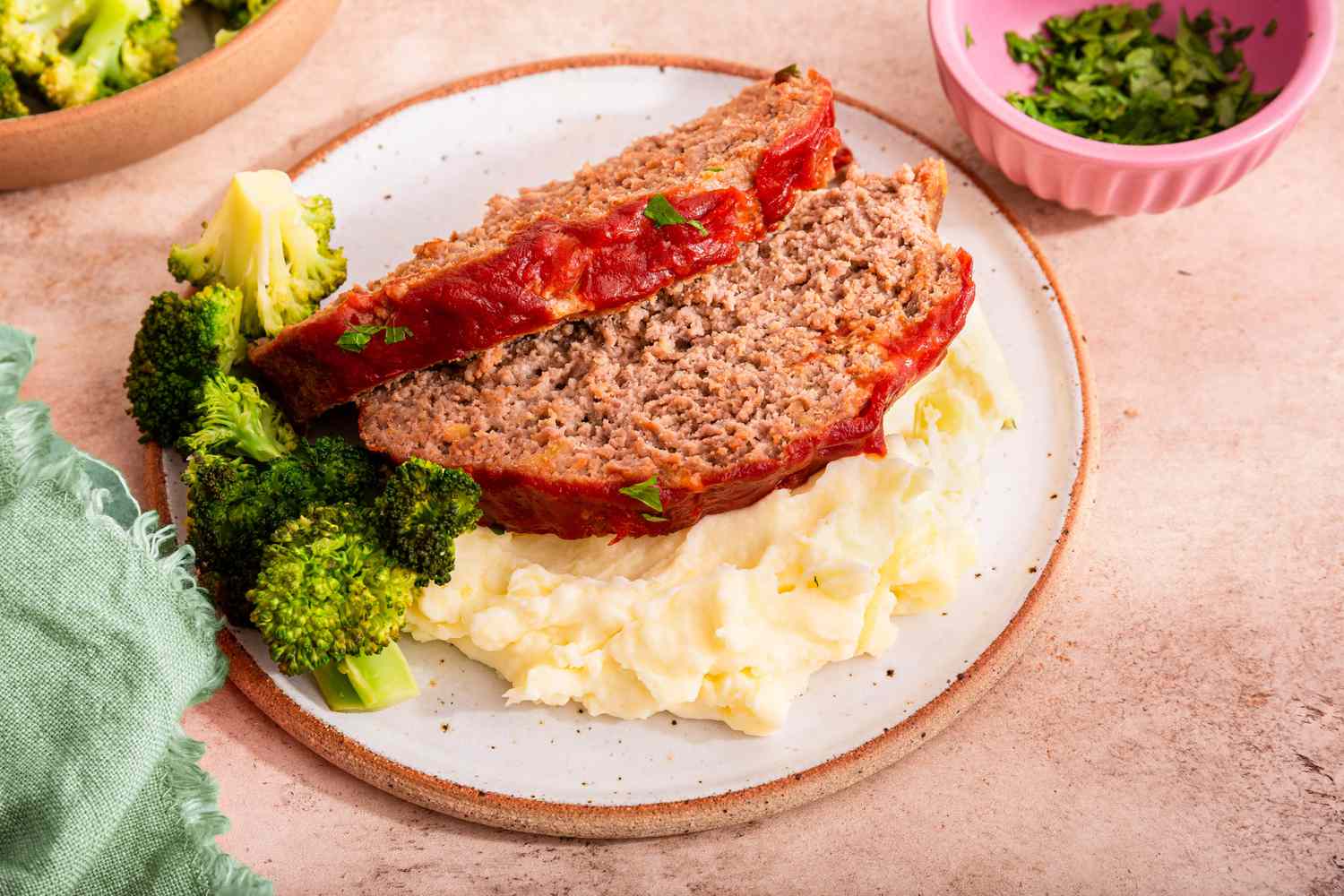 A Serving of Meatloaf, Mashed Potatoes, and Broccoli on a Plate Next to a Bowl With More Broccoli and a Bowl With Minced Parsley