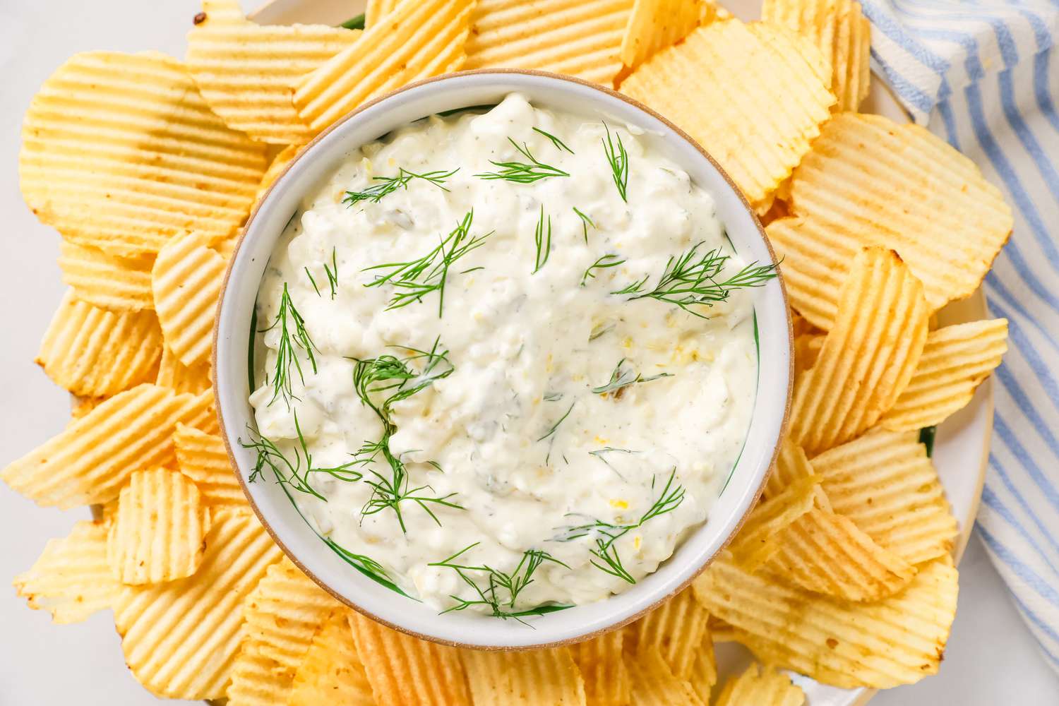 A bowl of creamy dip garnished with dill, surrounded by ridged potato chips