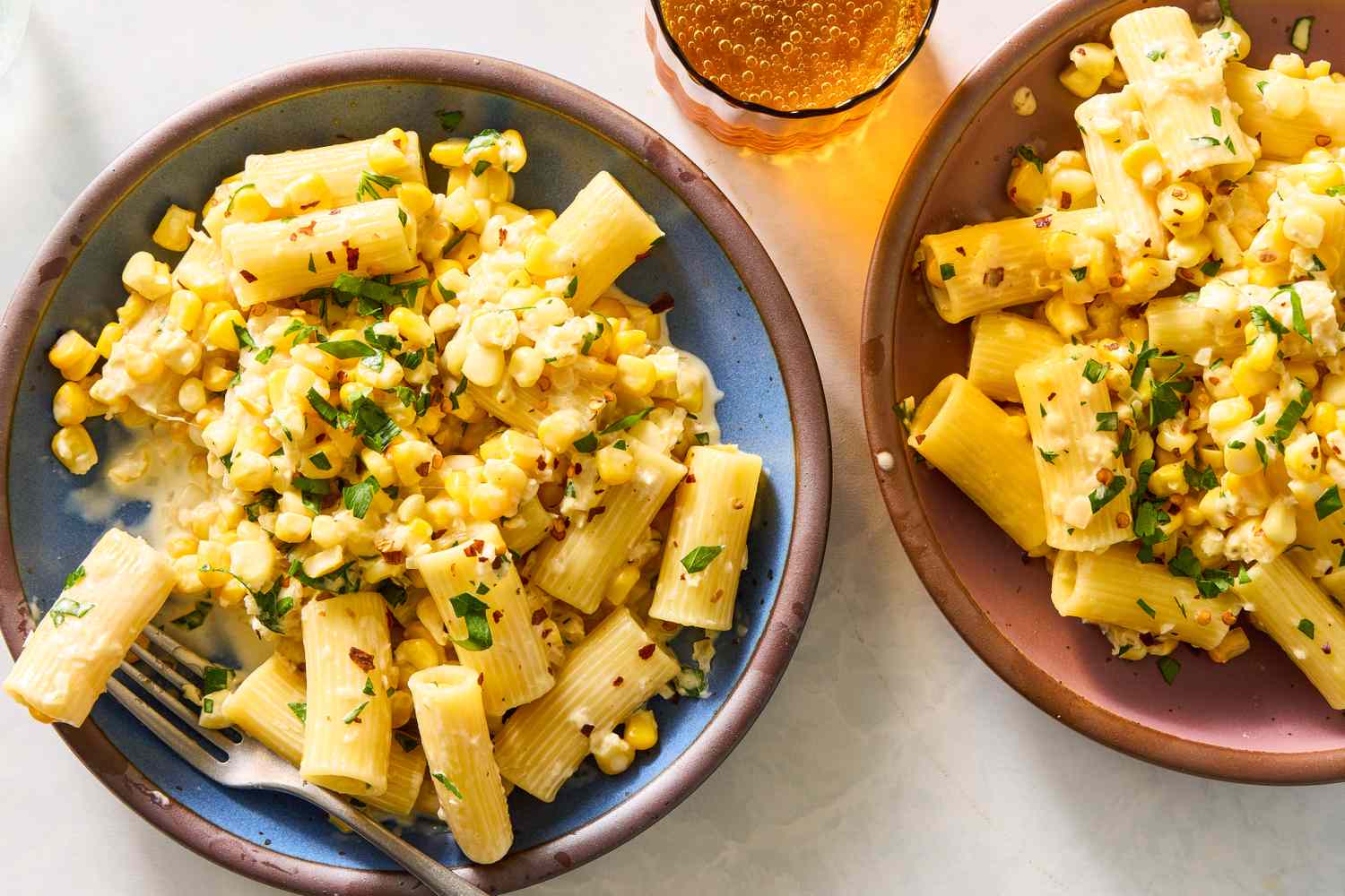 Two plates of pasta topped with corn and herbs next to a beverage in a glass