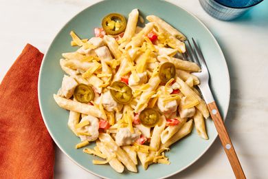Overhead view of rattlesnake pasta topped cheese and jalapenos light in green plate along with a fork and next to an orange napkin and blue drinking glass on a beige background