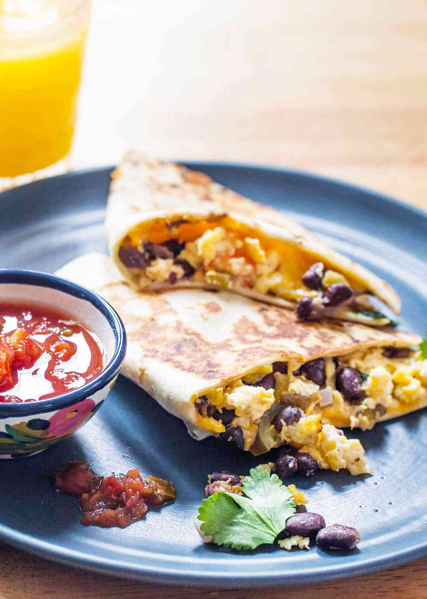 Side view of make-ahead breakfast quesadillas with a golden tortilla and the scrambled eggs, cilantro and black bean filling visible. A partial view of a glass of orange juice is above the blue plate to the left and a small bowl of salsa is next to the quesadillas on the plate.
