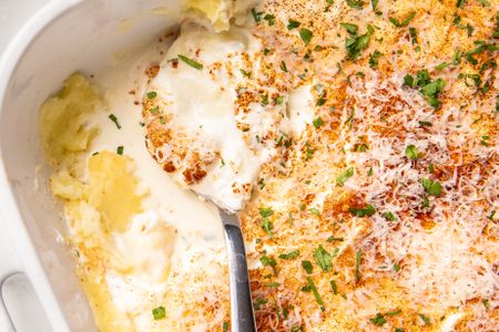 Casserole dish with a creamy layered casserole topped with grated cheese and parsley part of the dish served with a spoon