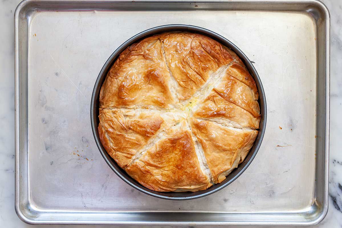 Overhead view of a cheese pie with artichokes set on a baking sheet. The top crust is golden and pale lines separate the slices on the crust.
