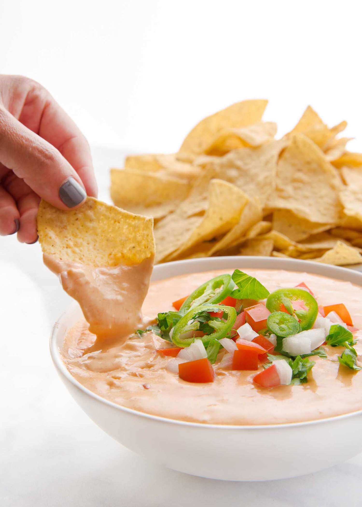 Hand scooping queso dip with pico de gallo and a bowl of tortilla chips nearby.