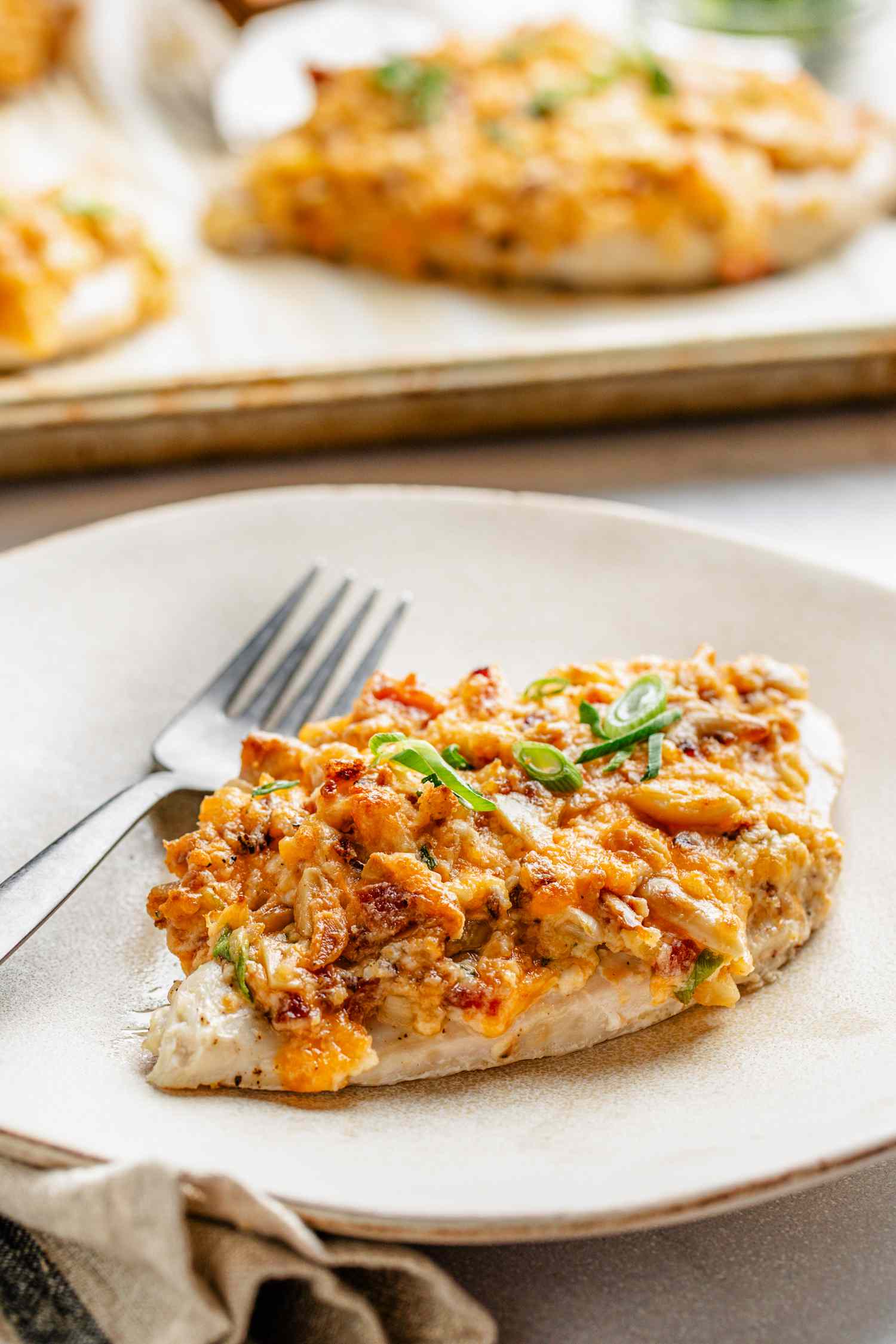 A million dollar chicken breast on a plate with a fork, and in the surroundings, a parchment paper lined baking tray with more