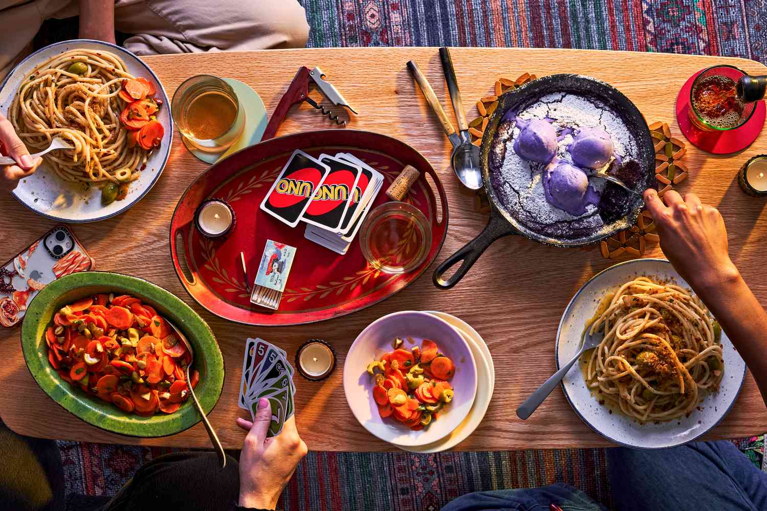 Coffee table filled with food including pasta, carrot salad, and ube skillet cookie, Hands are digging into the food and playing uno