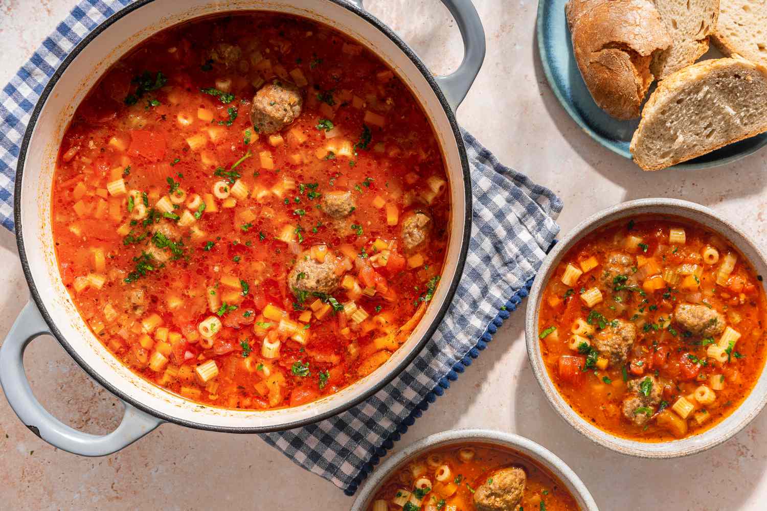 Overhead view of a pot of Italian meatball soup on a checkered towel next to two bowls of soup and a plate of bread