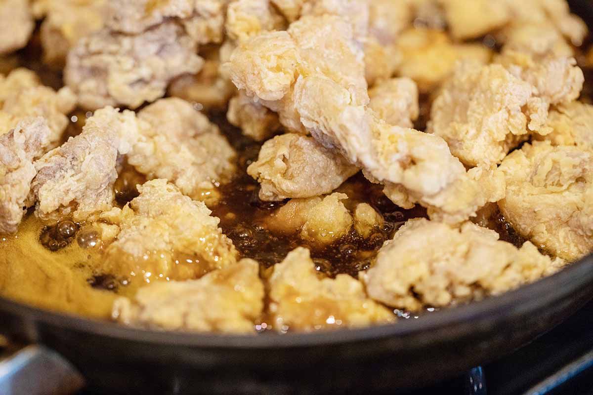 A cast iron skillet with fried chopped chicken on top of simmering orange chicken sauce.