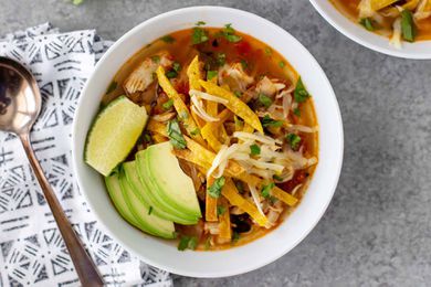 A white bowl of pressure cooker tortilla soup with a spoon and grey patterned linen napkin to the left. The top right corner has a partial view of a second bowl of the same soup.