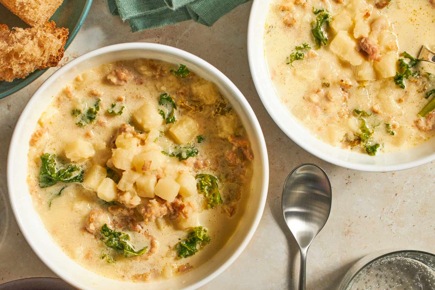 Two bowls of soup with sausage kale and potatoes accompanied by bread and a spoon