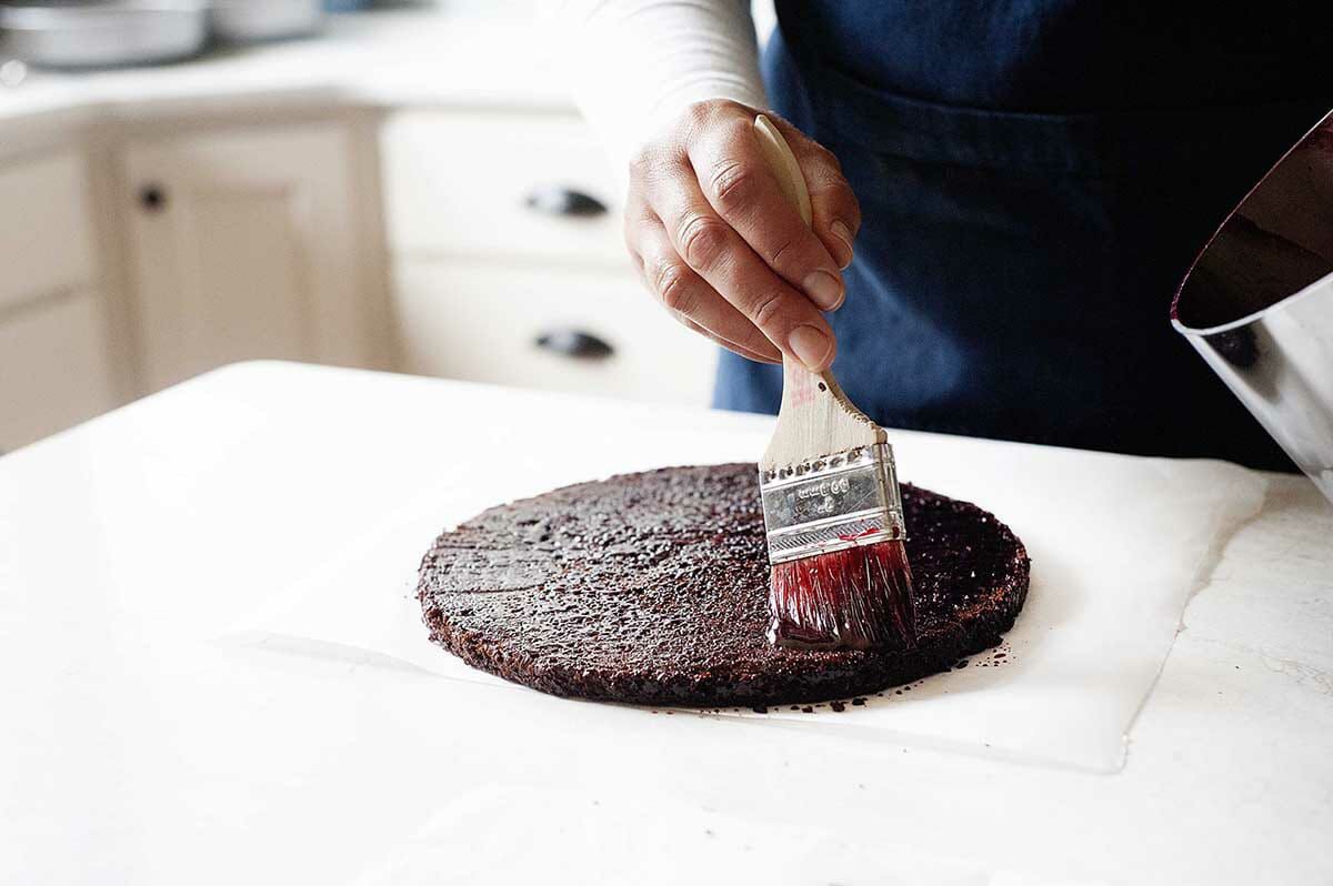 The first layer of chocolate cake is being brushed with cherry syrup to show how to make Black Forest Cake.