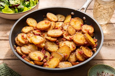 Panful of Bratkartoffeln (German Cottage Fries) at a rustic table setting with a bowl of salad, a glass of water, and a small saucer with salt and pepper