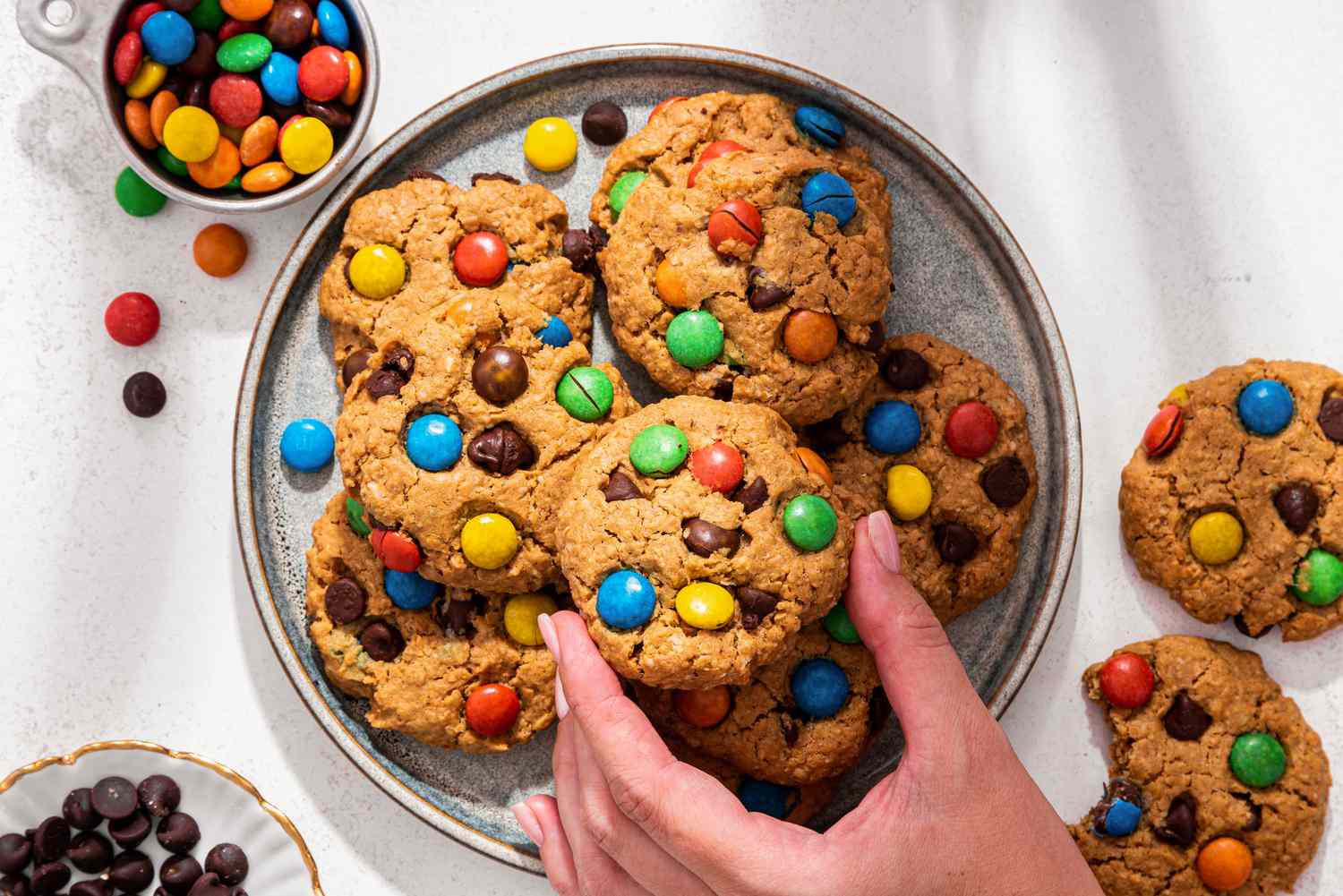 A plateful of monster cookies with chocolate chips and M&Ms on the side, and a hand reaching in to grab one