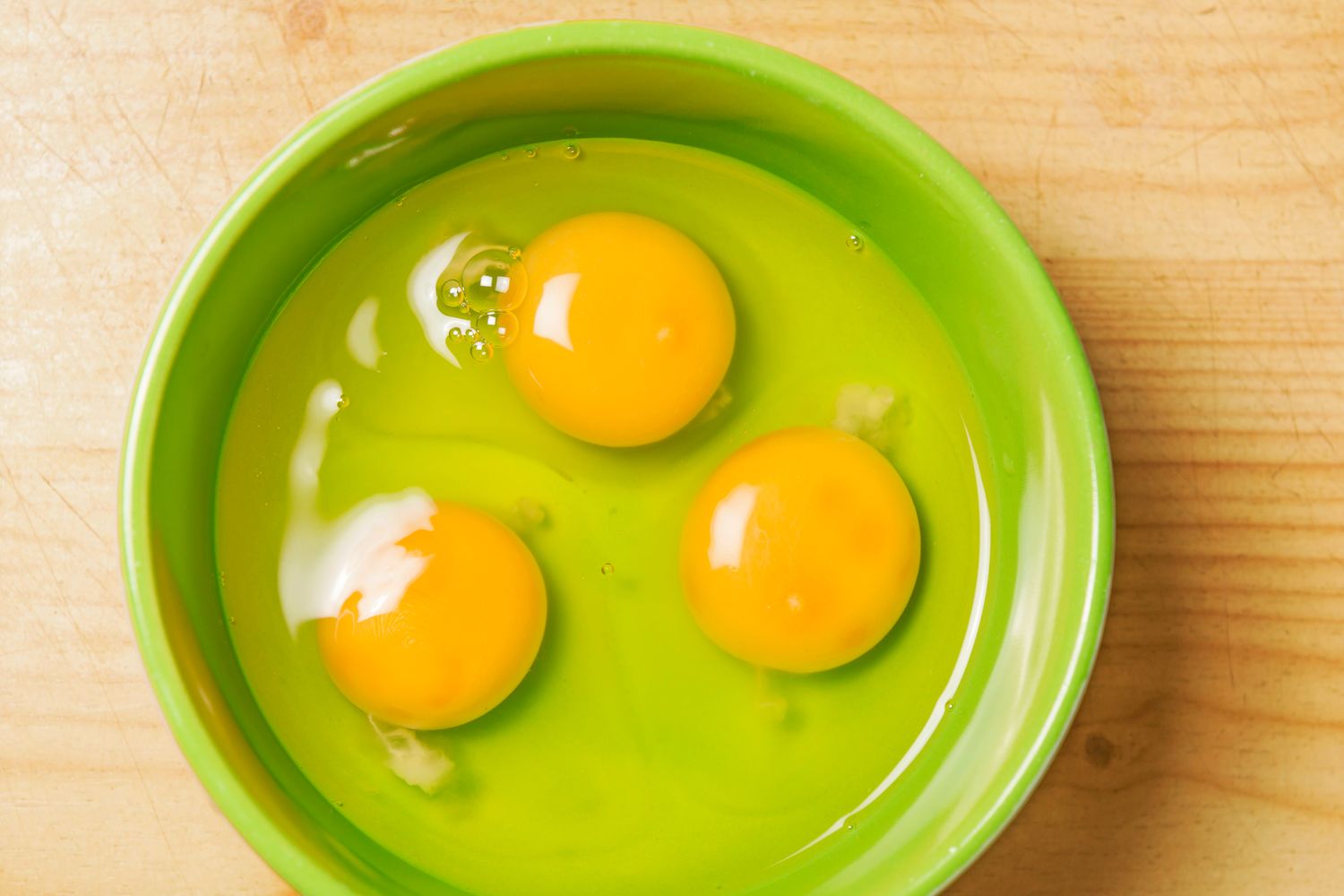 Three raw egg yolks in a green bowl on a wooden surface
