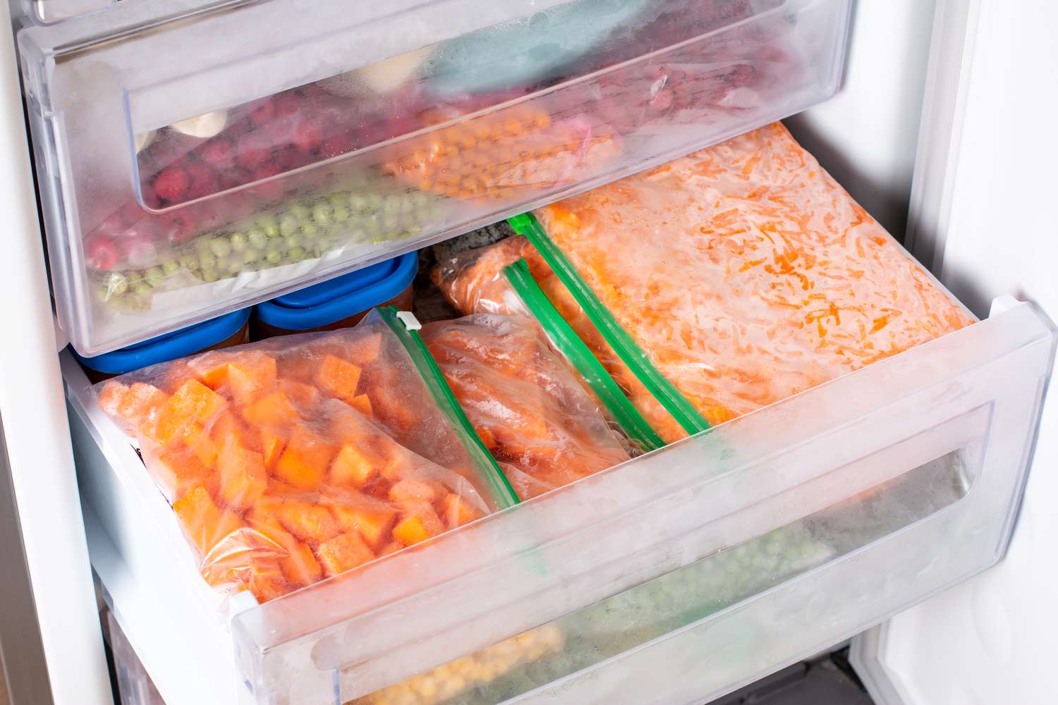 Angled view of one open and one closed freezer drawer stacked full of various frozen fruits and vegetables in plastic zipper style storage bags