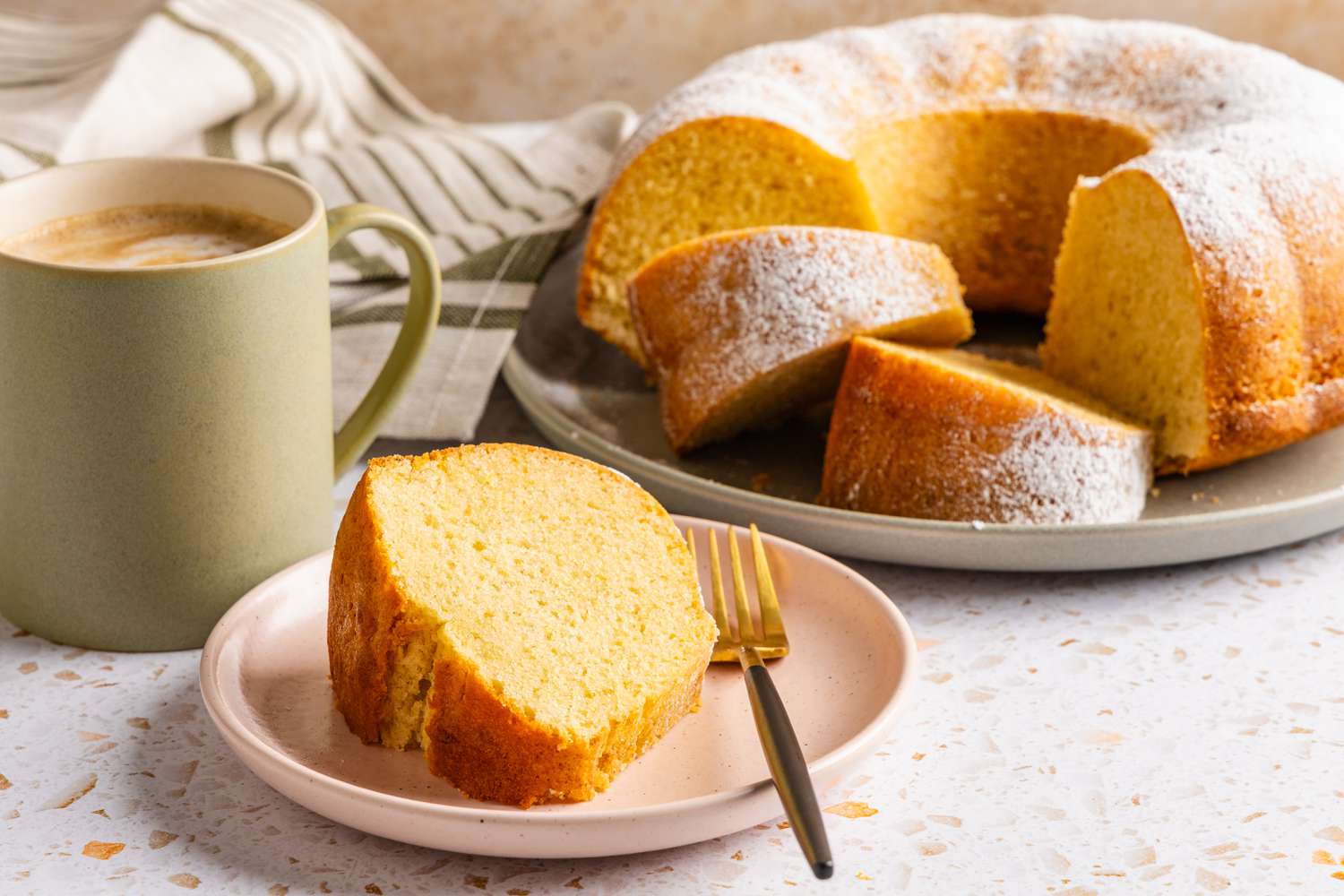slice of ciambellone on a plate with a fork, and in the background, a cup of coffee, more cake on a platter, and table napkins
