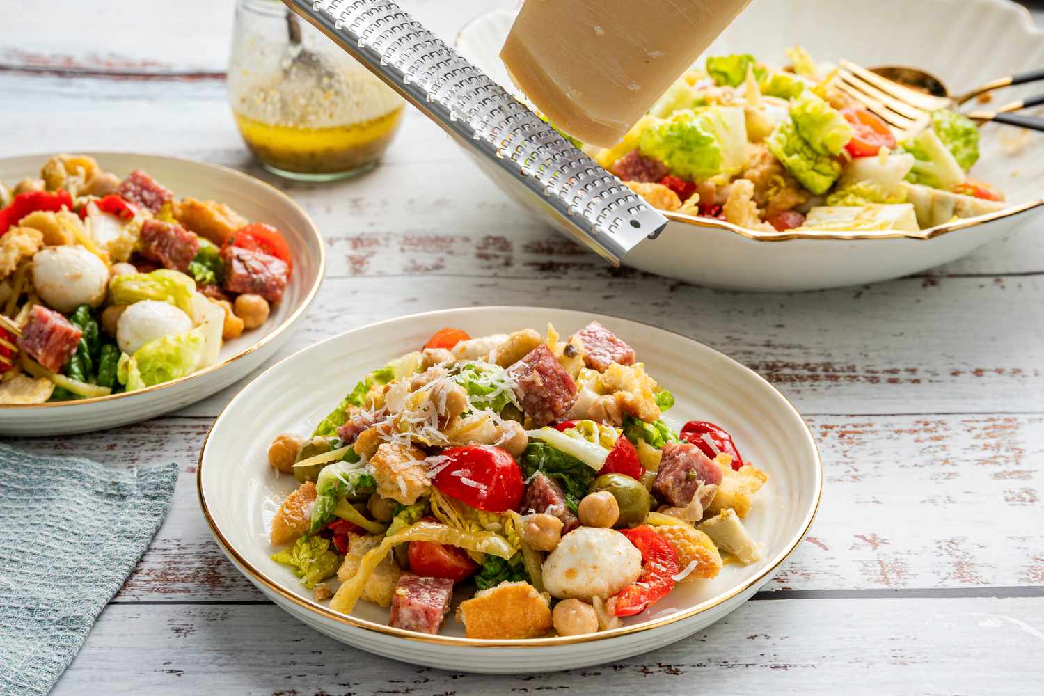 Fresh Parmesan Grated Onto a Serving of Antipasto Salad on a Plate Using a Microplane, and in the Surroundings, Another Serving on a Plate, a Bowl With the Rest of the Salad, and a Jar With More Dressing 