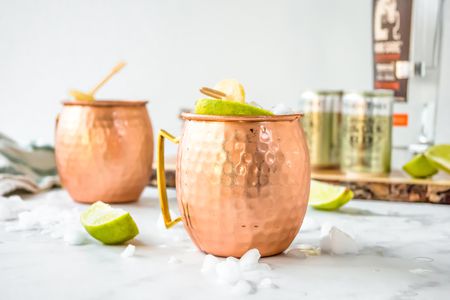 Mezcal Mule in Two Copper Mugs Surrounded by Ice and Limes. More Ingredients in the Background