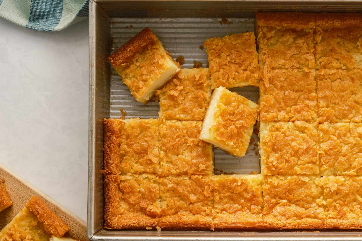 Overhead shot of butter mochi in a rectangular baking dish, cut into squares, with some pieces of mochi put on a wooden board on the side of the baking dish