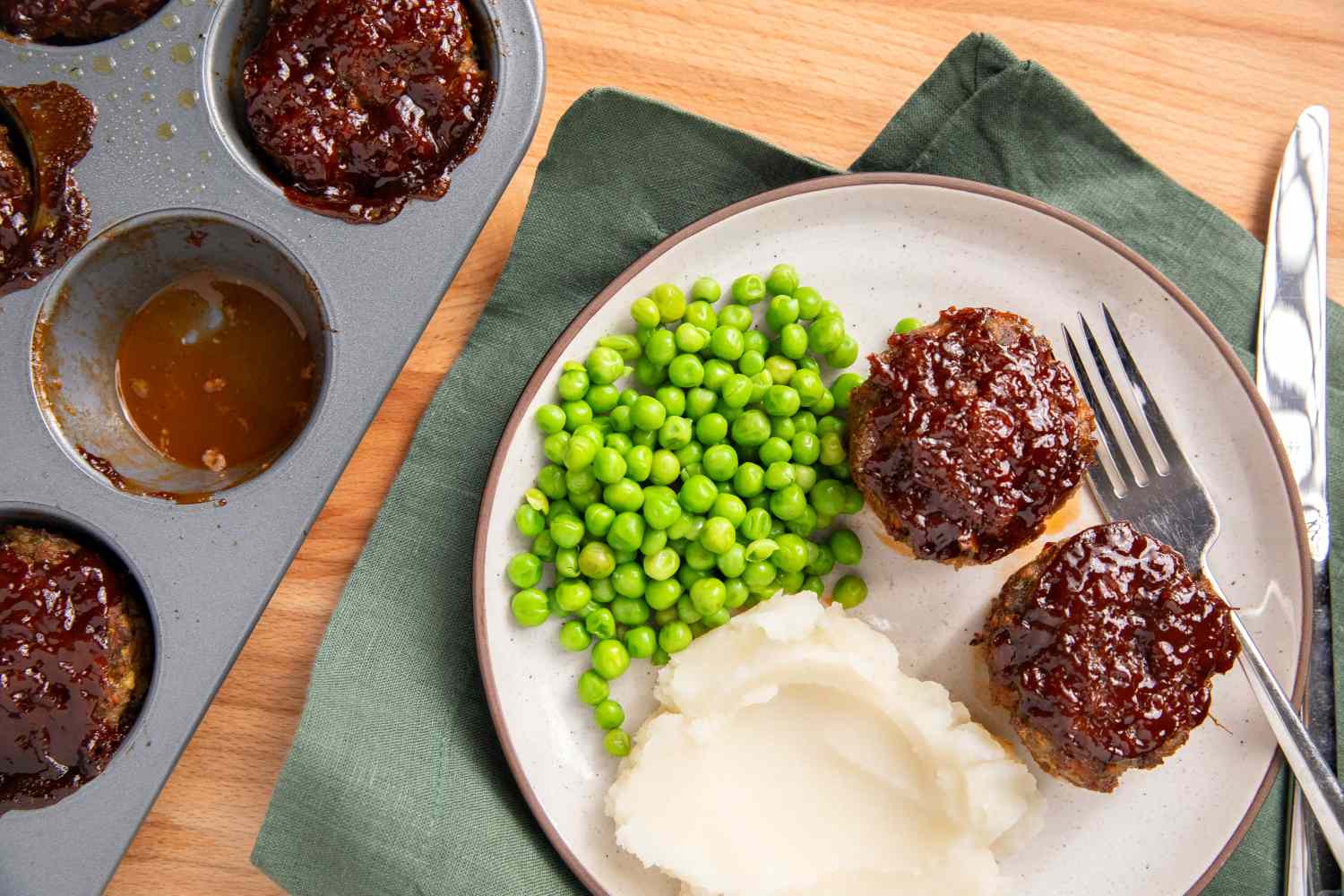 A plate of meatloaf muffins with mashed potatoes and peas next to a muffin tin with remaining muffins