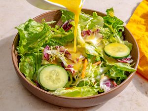 Angled view of pouring Miso-Maple Dressing over lettuce and cucumbers in a brown bowl all on a tan countertop