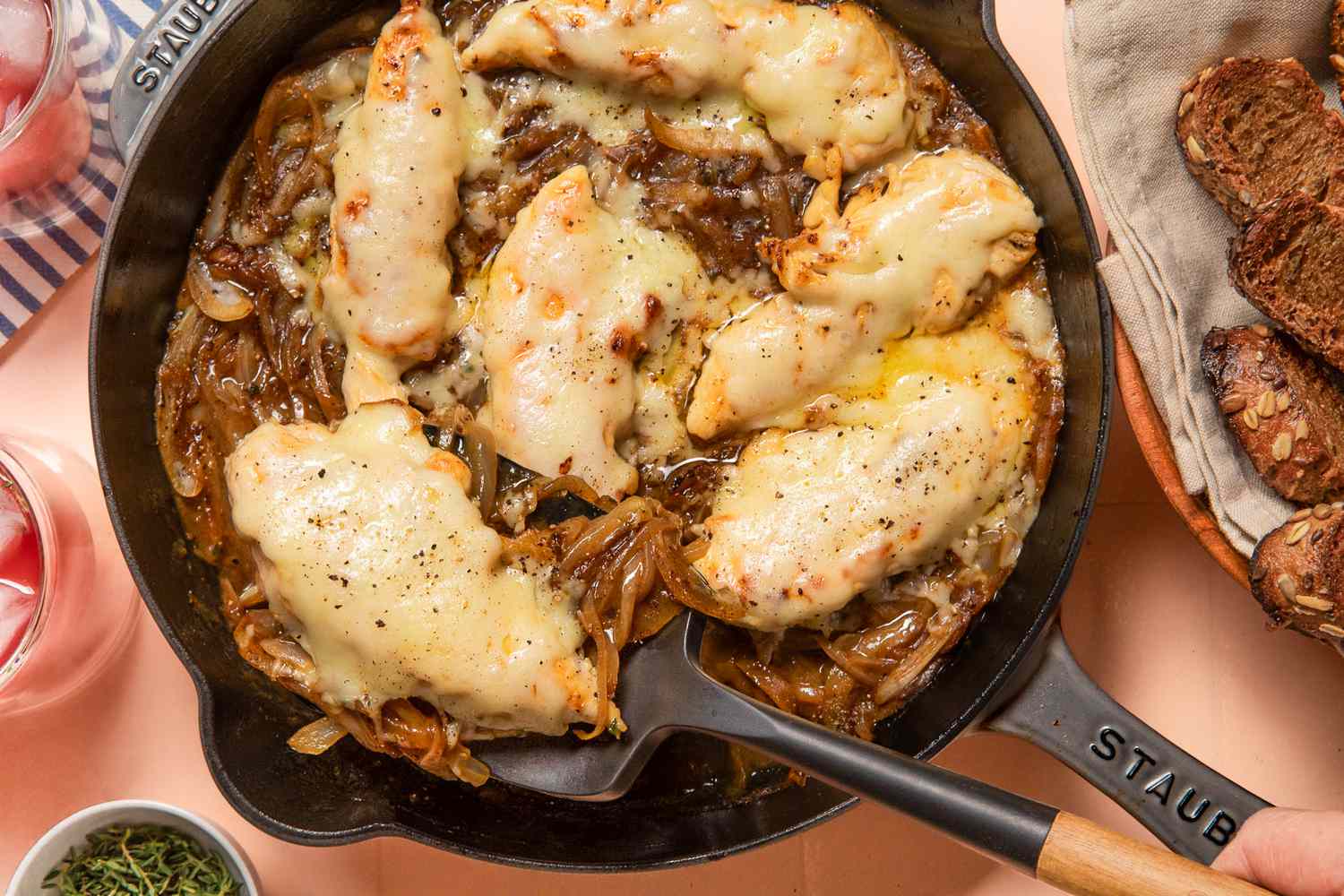 french onion chicken in a skillet at a table setting with glasses, a bowl of bread, and a small bowl of herbs