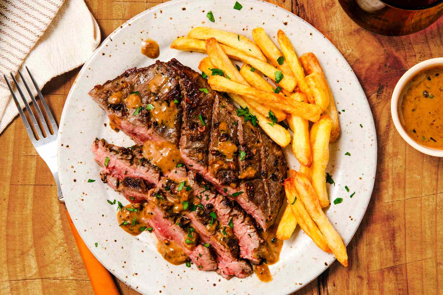 Overhead view of a platter of partially sliced flank steak and french fries next to a small bowl of sauce and fork all on a wooden tabletop