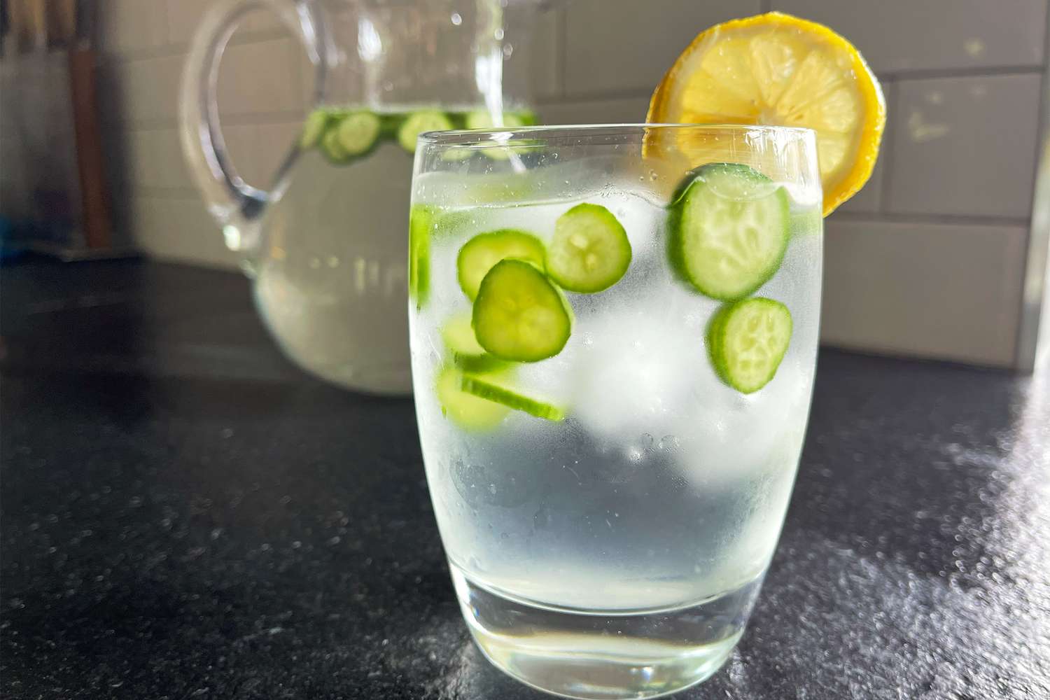 Glass of water with cucumber slices and a lemon garnish, pitcher of infused water in the background
