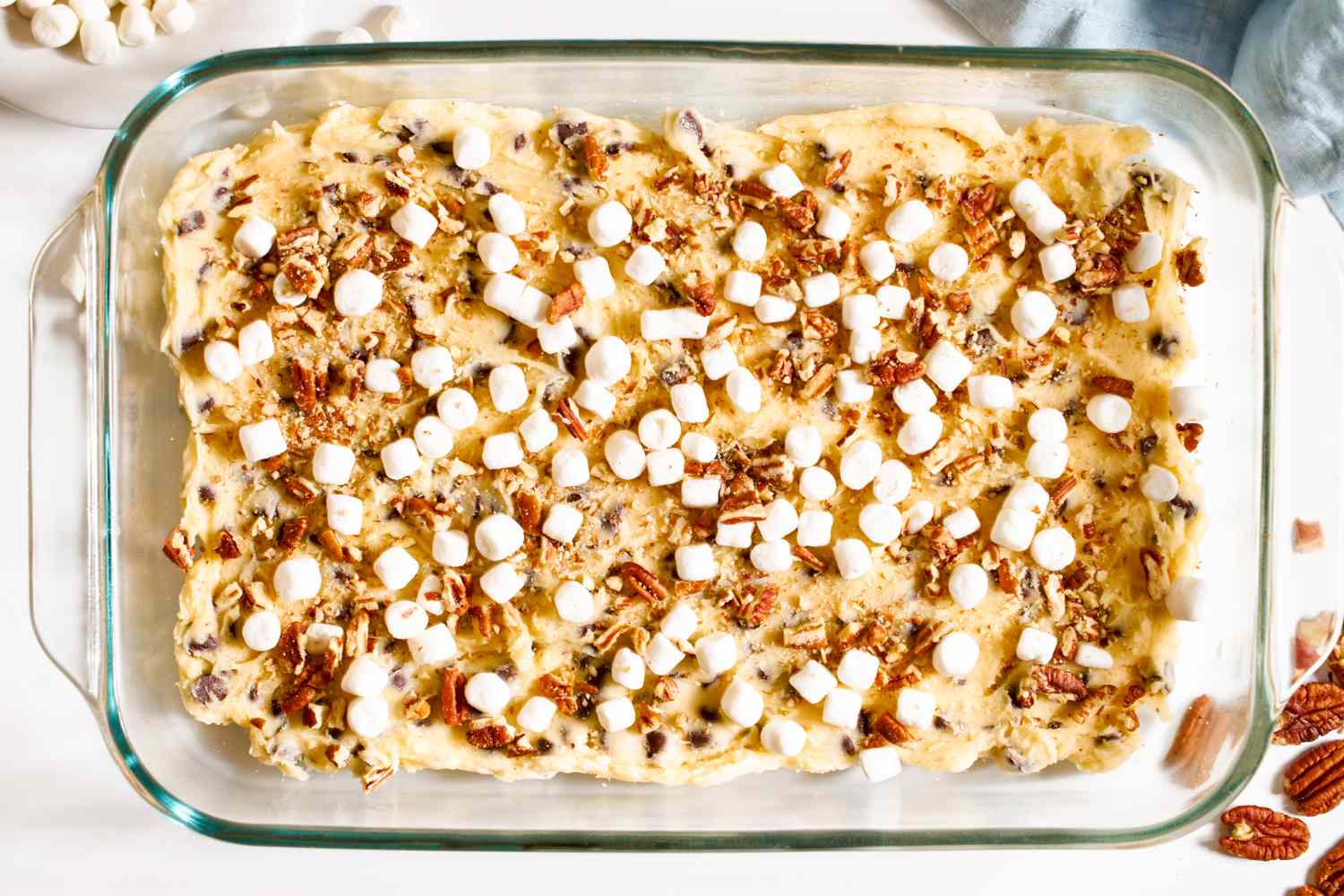 Cookie dough in square glass balking dish topped with mini marshmallows and chopped pecans for mud hen bars recipe