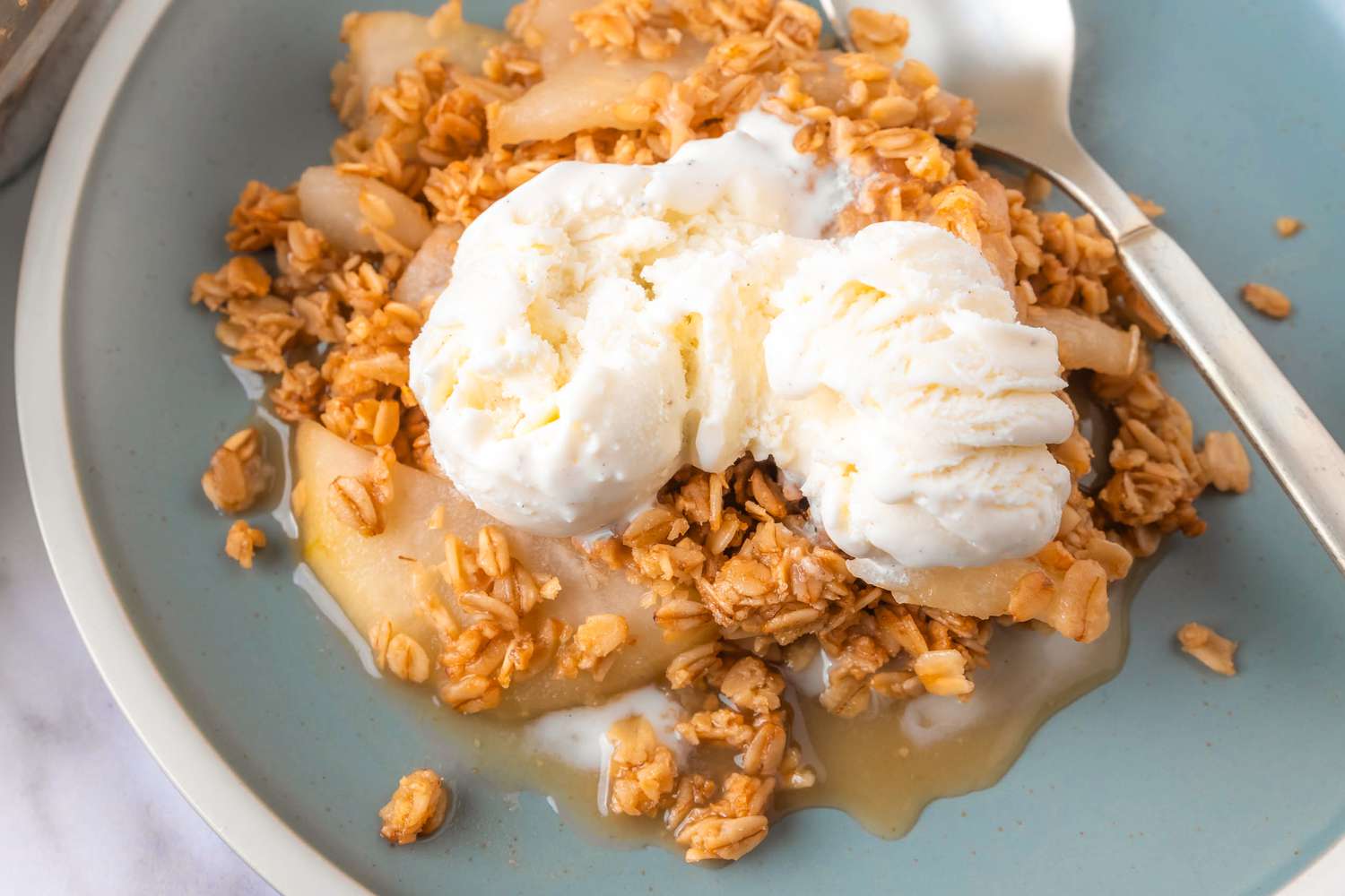 Overhead view of a teal plate of vanilla pear crisp recipe topped with vanilla ice cream and next to a spoon