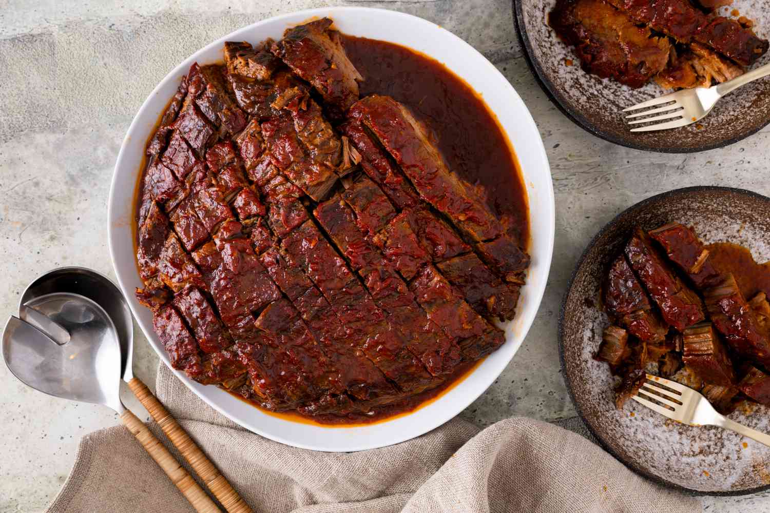 Overhead view of a white platter of sliced beef brisket next to serving spoons and two plates of brisket with forks all on a marbled countertop