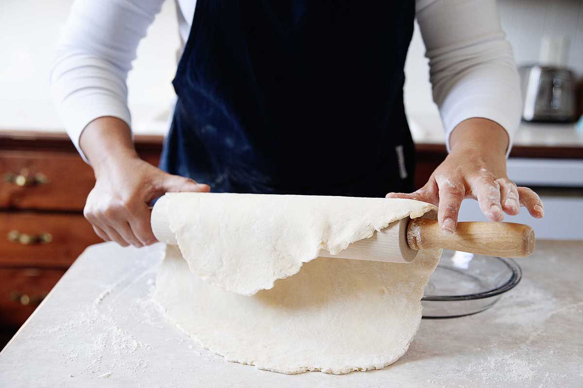 Woman lifting pie crust off of the counter with a rolling pin and placing it into a pie plate.