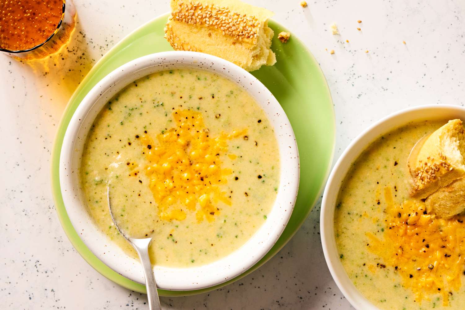 Overhead view of a two bowls of broccoli cheddar soup, one with a spoon and on a plate with pieces of bread, one with a piece of bread in the soup all next to a drink glass