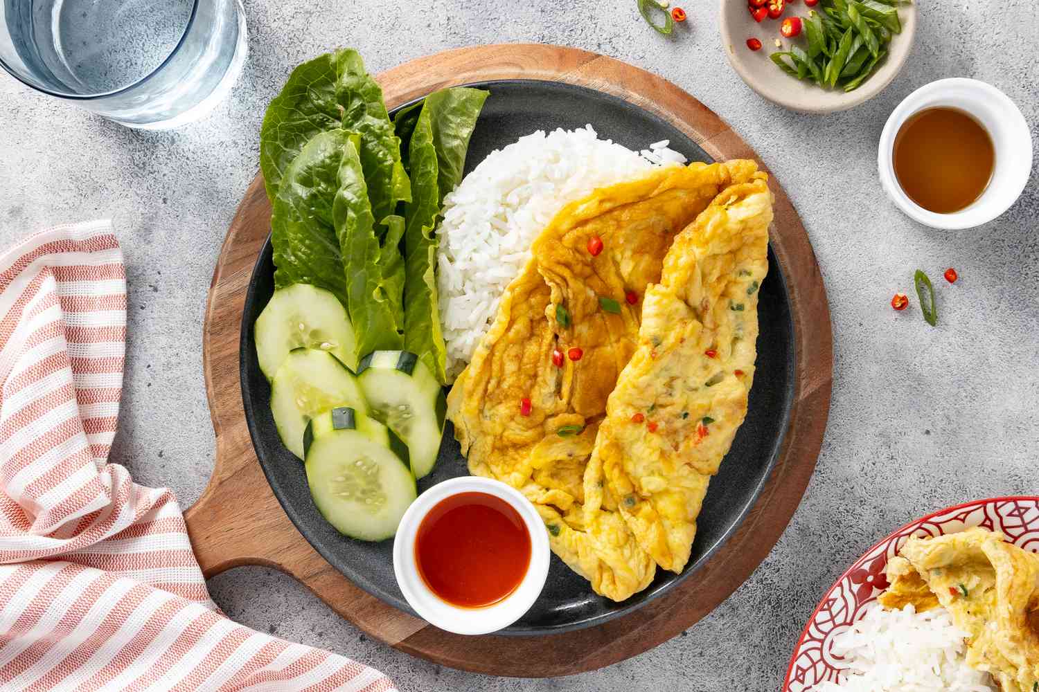 Plate of Kai Jeow (Thai Omelet) with Cucumber Slices, Romaine Leaves, Rice, and a Small Bowl of Sriracha on a Circular Wooden Serving Board and Surrounded by a Table Setting with a Glass of Water, a Table Napkin, a Small Bowl of Slice Scallions and Thai Chili, a Small Bowl of Fish Sauce, and Another Plate of Kai Jeow