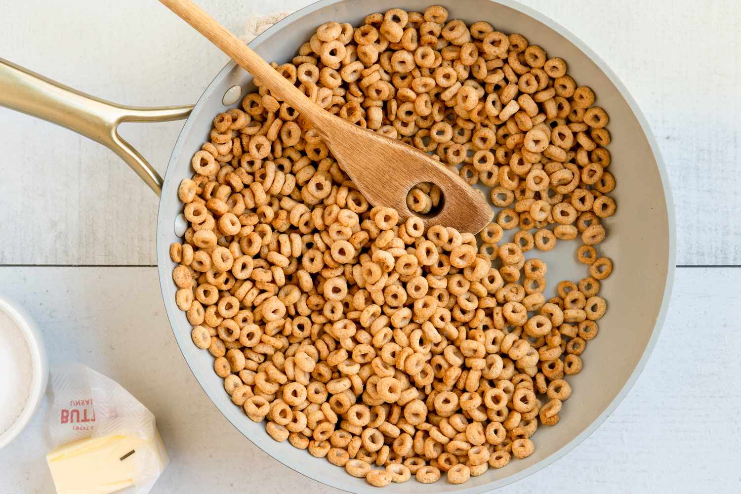 A skillet with Cheerios being stirred by a wooden spoon a pat of butter is nearby