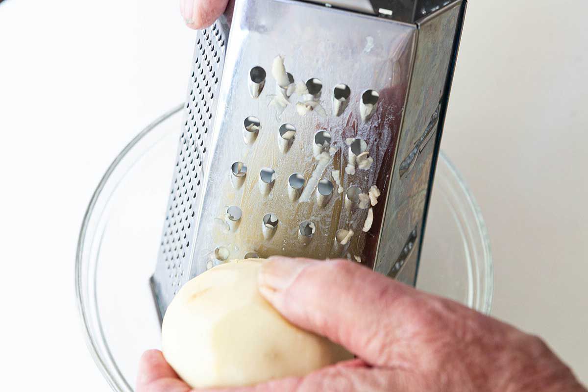 A potato being grated into a glass bowl 