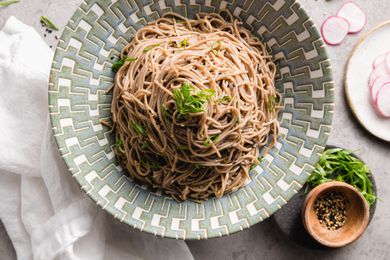 Sesame Soba Noodles in a Bowl Garnished with Spring Onions Next to Plate with Spring Onions and Seasoning and Another Plate with Slices of Radish
