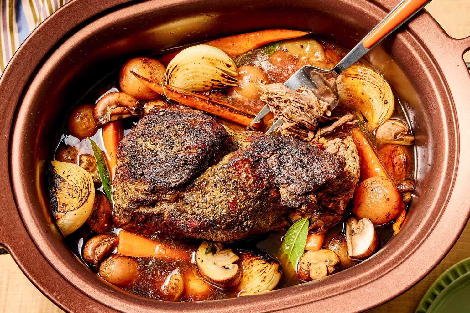 A slowcooked pot roast with vegetables in a crockpot shown with a fork lifting a piece of the roast