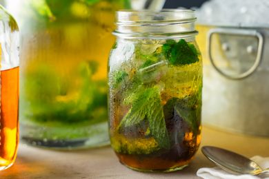Side view of a mason jar of mint julep, ice and mint leaves with a tub of ice and pitcher in the background