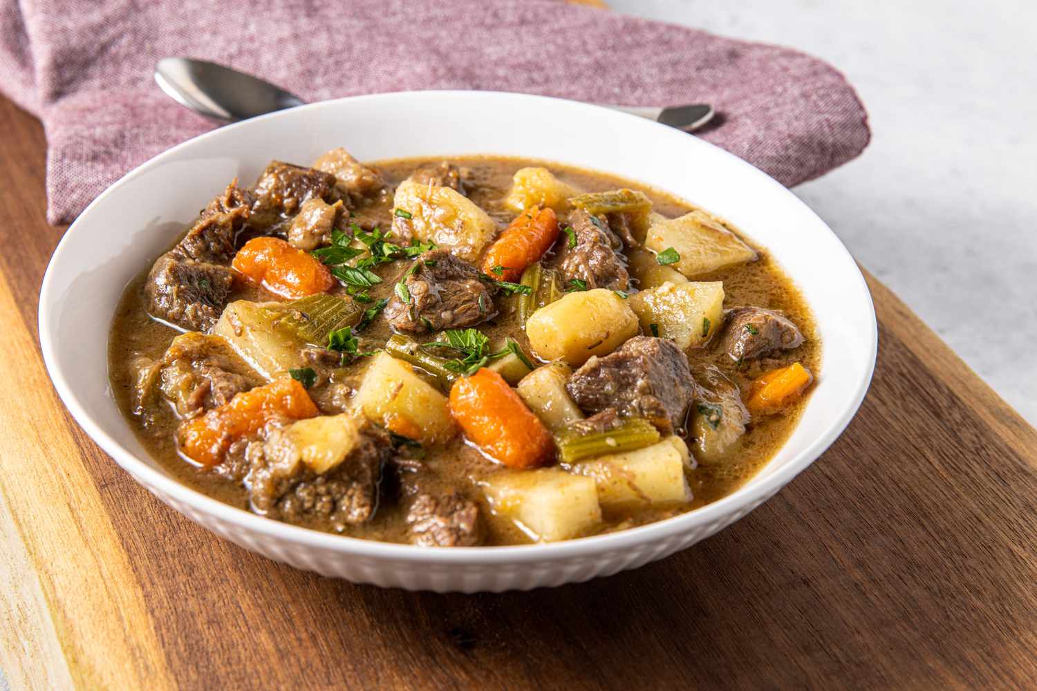 Bowl of Easy Beef Stew Toped with Some Parsley and in the Background, a Kitchen Towel with a Spoon on Top
