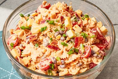 A bowl of pasta salad with tomatoes, beans, cheese, green onions, and bacon bits, on a table